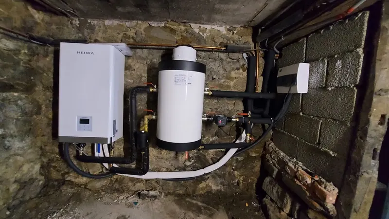 Chauffe-eau compact neuf raccordé avec tuyaux isolés noirs dans une cave en pierre, vue large d'un remplacement réalisé par AS Depann