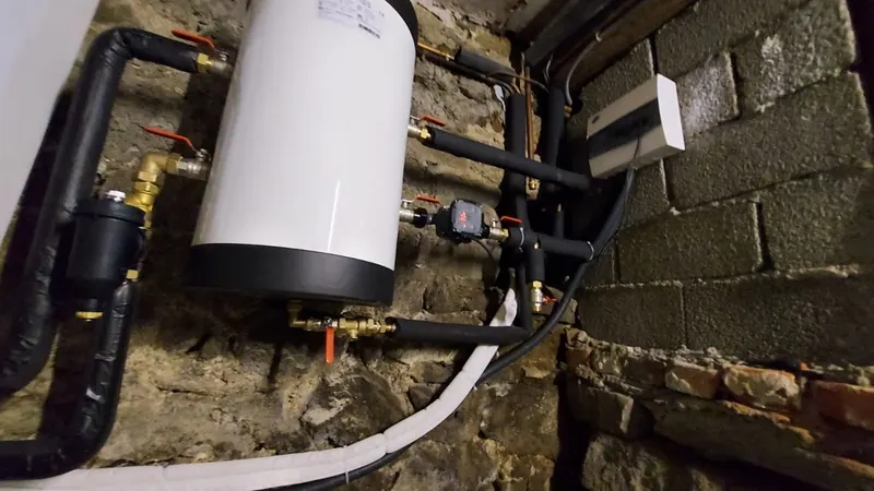 Chauffe-eau électrique neuf raccordé avec tuyaux isolés noirs contre un mur en pierre de cave, pose AS Depann