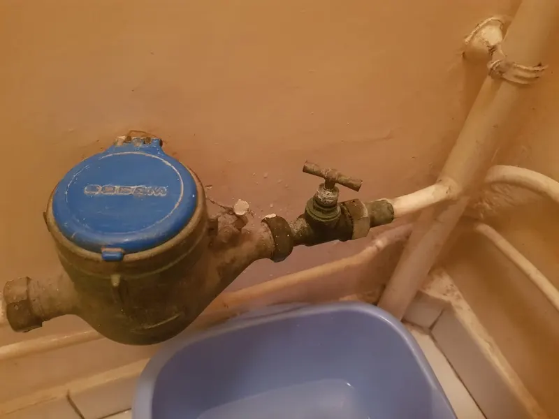 Compteur d'eau bleu ancien et robinet d'arrêt corrodé au-dessus d'une bassine, compteur mécanique ancien avec ses vannes, lecture décrite dans le guide AS Depann