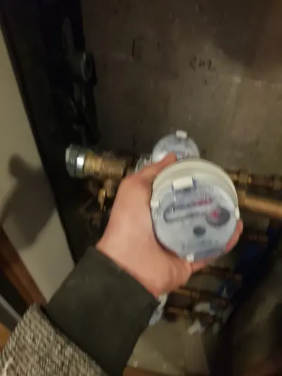 Compteur d'eau tenu en main devant une nourrice avec vannes laiton, cadran avec chiffres noirs et rouges et petite roue, lecture expliquée dans le guide AS Depann