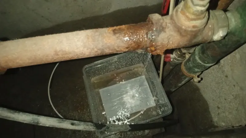 Bac en plastique recueillant les gouttes sous une canalisation en fonte rouillée qui fuit, réseau ancien tel que AS Depann en répare dans les immeubles lyonnais d'avant-guerre