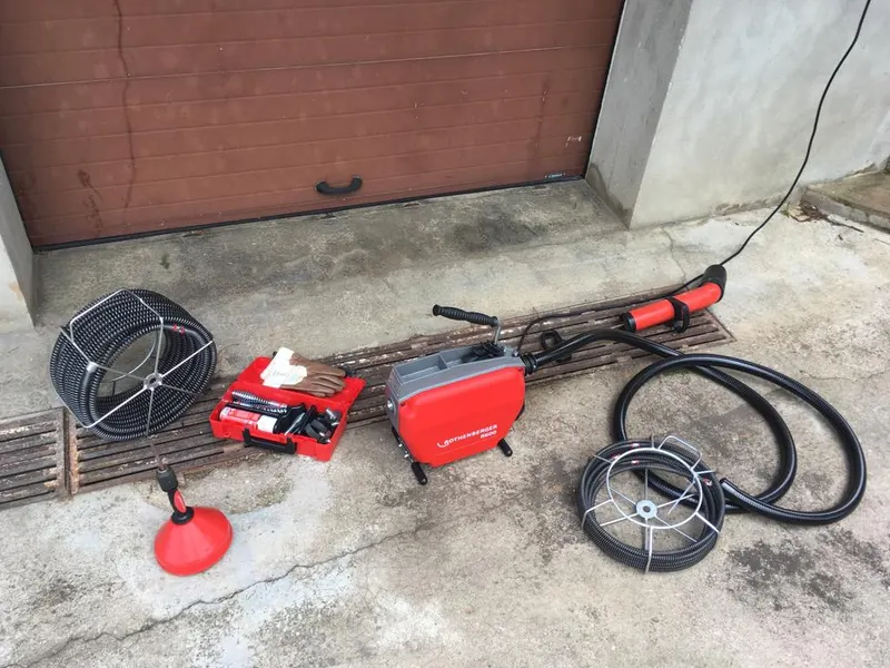 Furet électrique rouge et flexibles de débouchage posés devant une porte de garage avant intervention, matériel de AS Depann pour un regard bouché dans une maison de Millery
