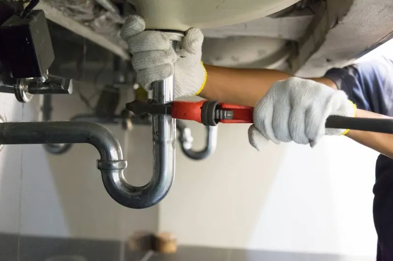 Plombier serrant un siphon chromé sous un lavabo avec une pince, réparation type d'une fuite sous évier (photo d'illustration)